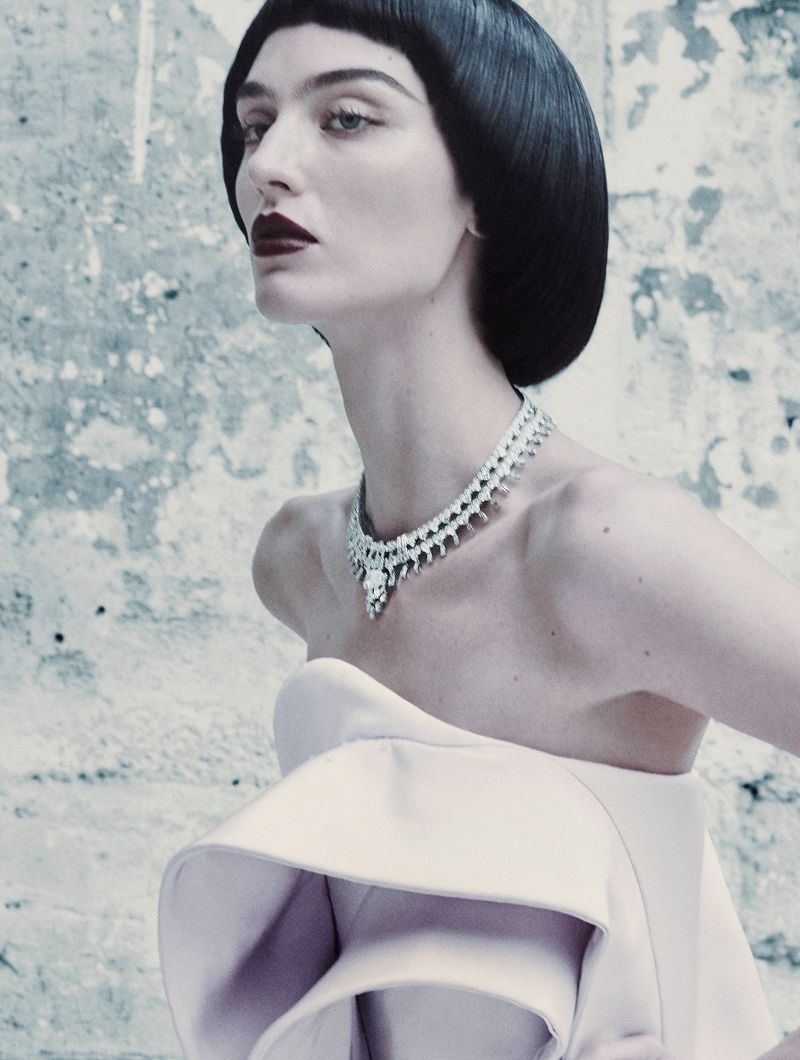 Fashion portrait of a woman with dark bob haircut wearing a diamond necklace and strapless dress against a textured grey background.