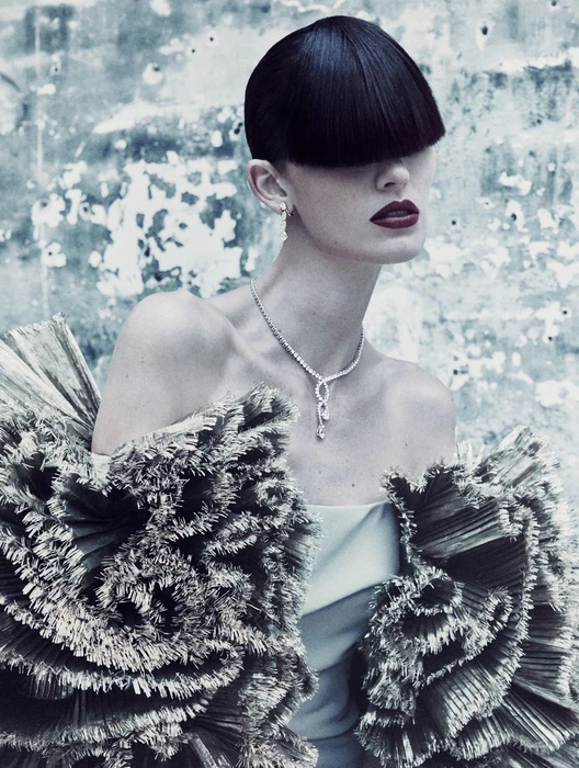 Model with heavy blunt fringe wearing a textured fringe garment and diamond necklace against a distressed wall.