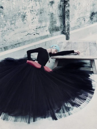 Model in a black long-sleeve top and large tulle skirt reclining near a wooden table against a weathered concrete wall
