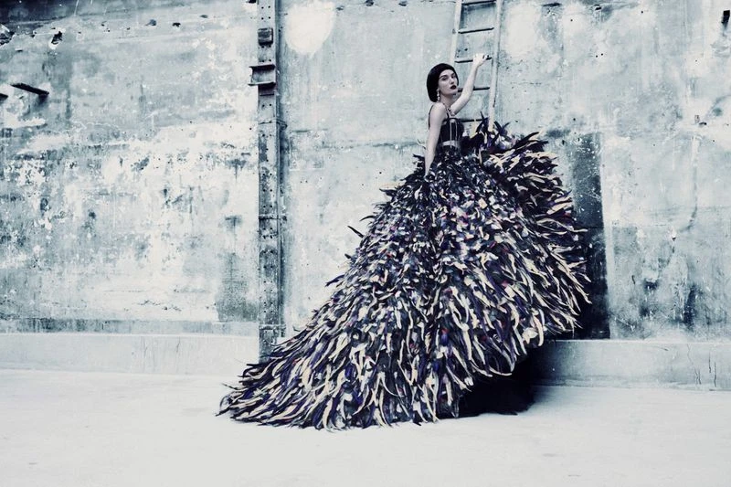 Model in voluminous feathered dress standing against an industrial textured wall with a ladder