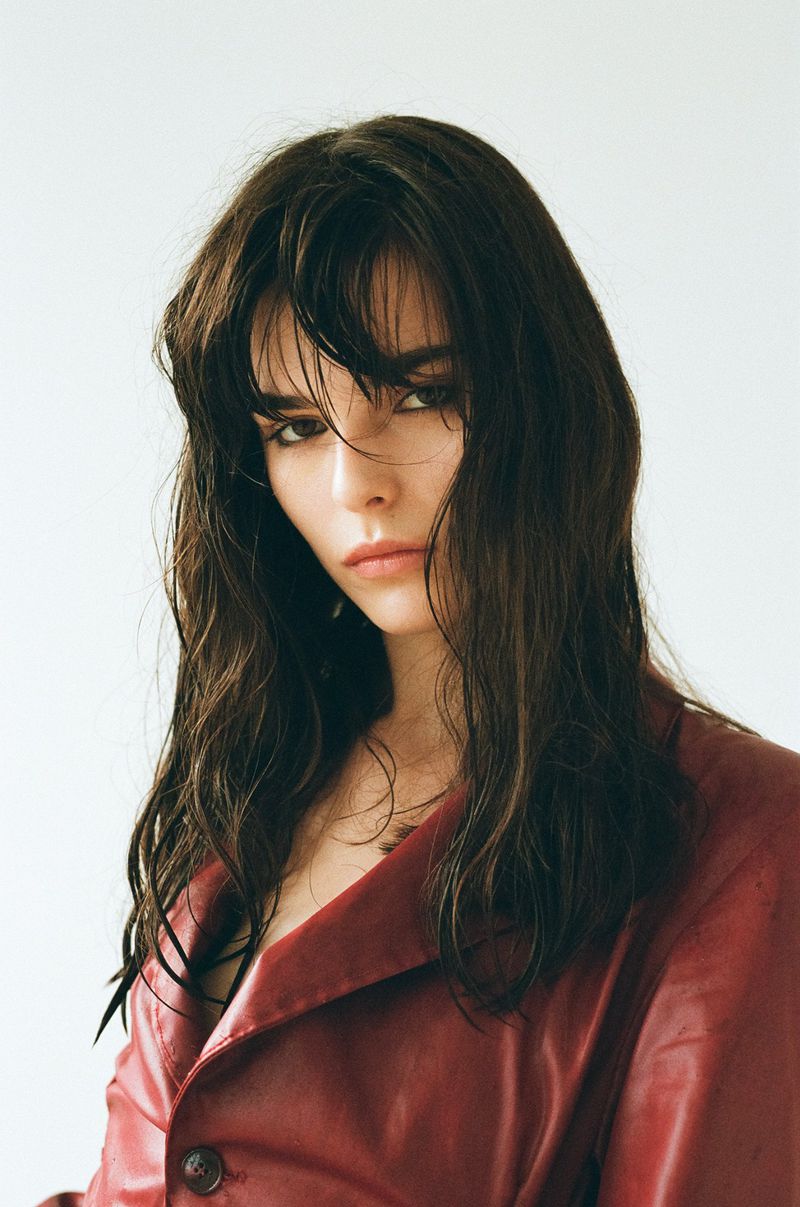 Close-up studio portrait of a young adult woman with long, dark, wavy hair, wearing a red coat against a plain white background.