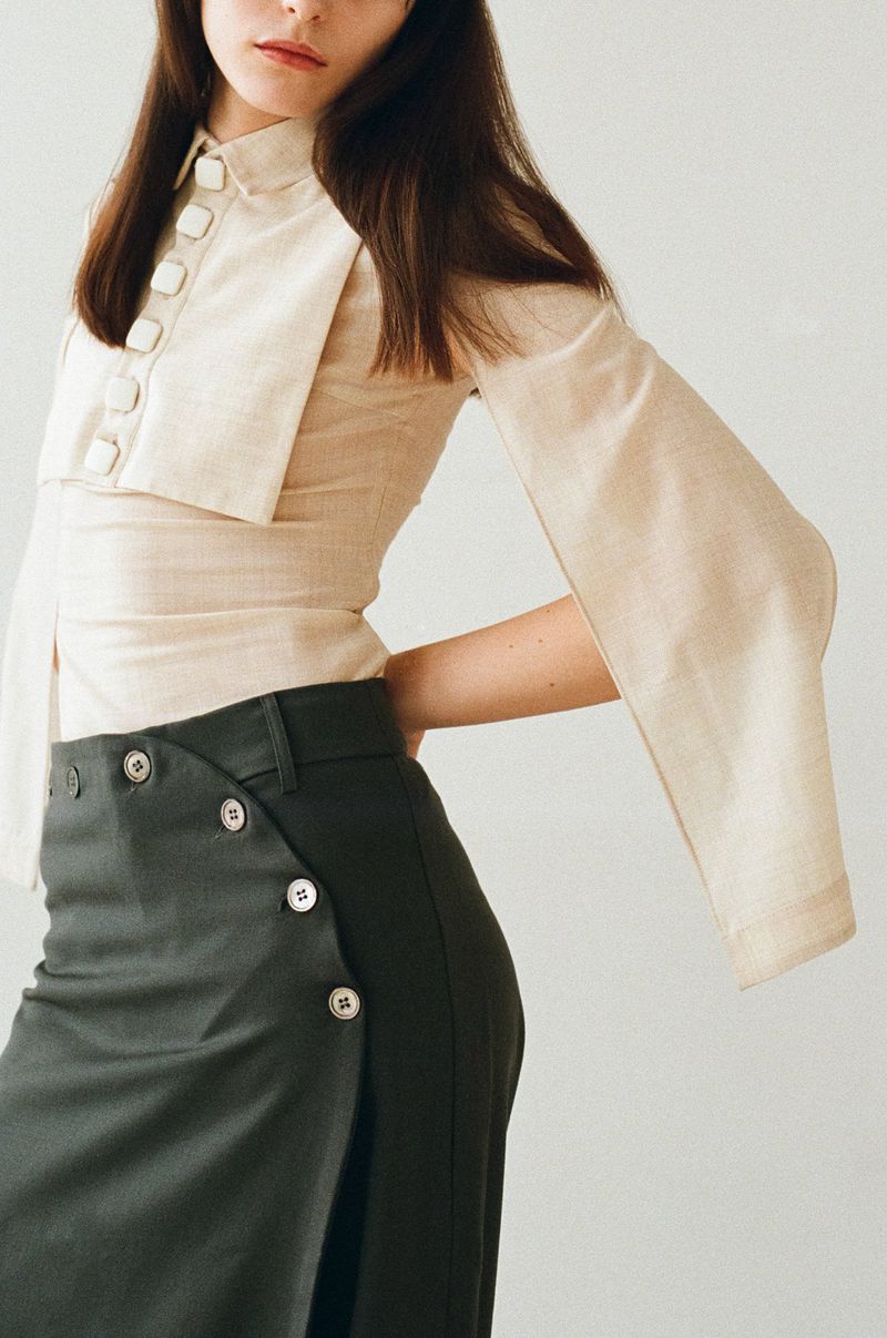 Brunette woman wears a cream-colored shirt and dark green skirt in a minimalist studio fashion shot. Natural lighting highlights the fabric.