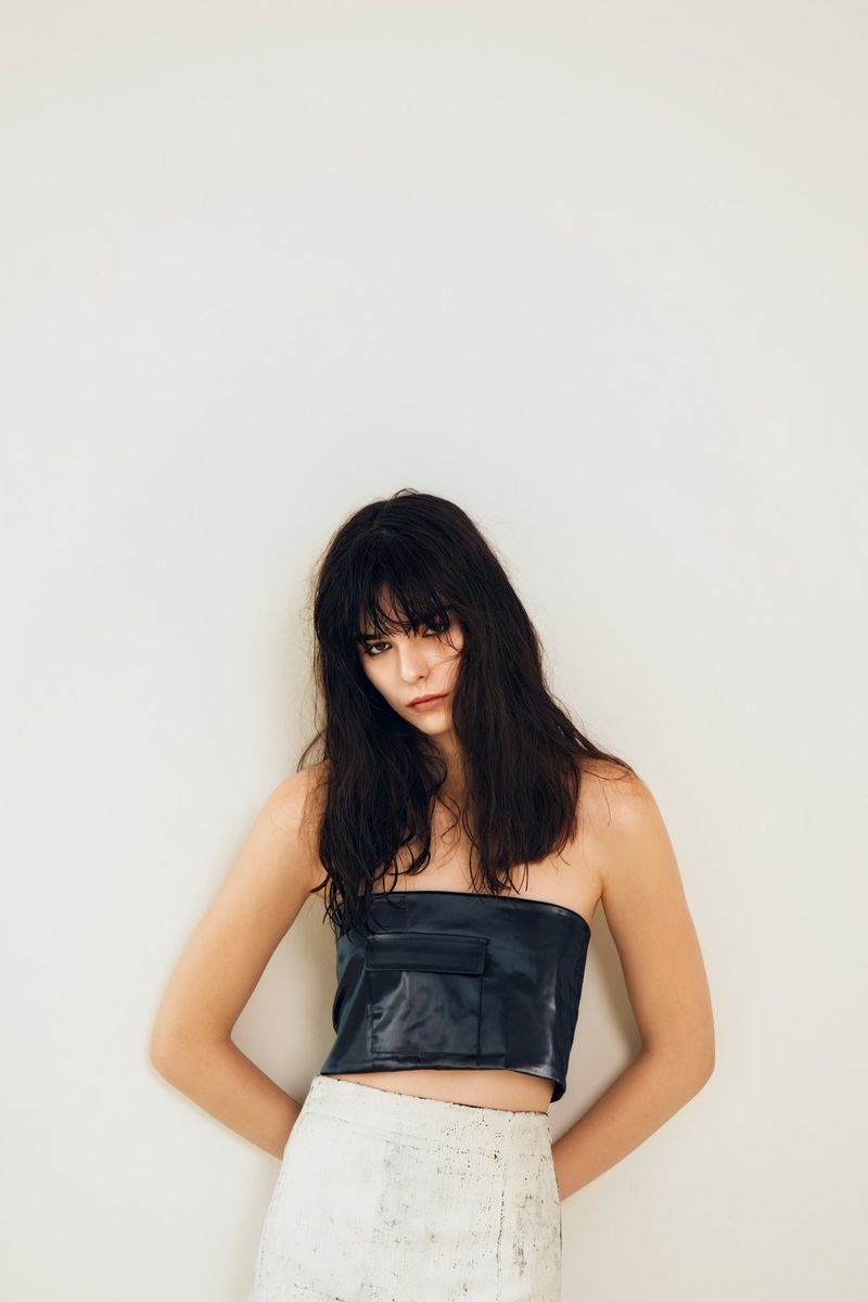 Fashion shot of a young adult woman in a dark, strapless top and white skirt, hands behind her back, standing against a neutral wall.