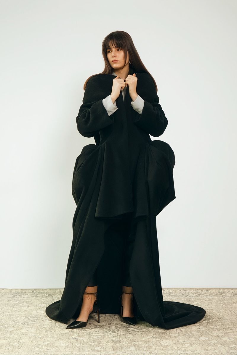 A woman posing indoors with a neutral background, wearing a dramatic and voluminous black coat with heels, in a fashion editorial style.