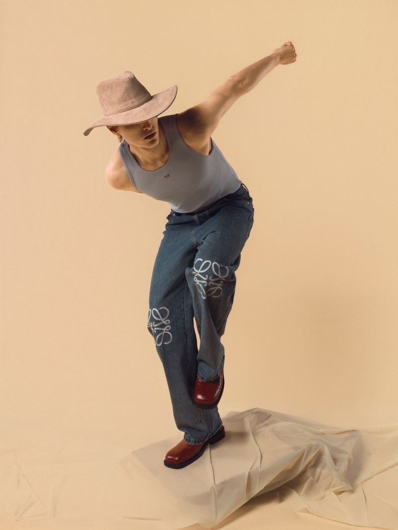 Fashion shot of a male model in a light blue tank top, jeans with Loewe logo, and a cowboy hat, posing on a beige backdrop.