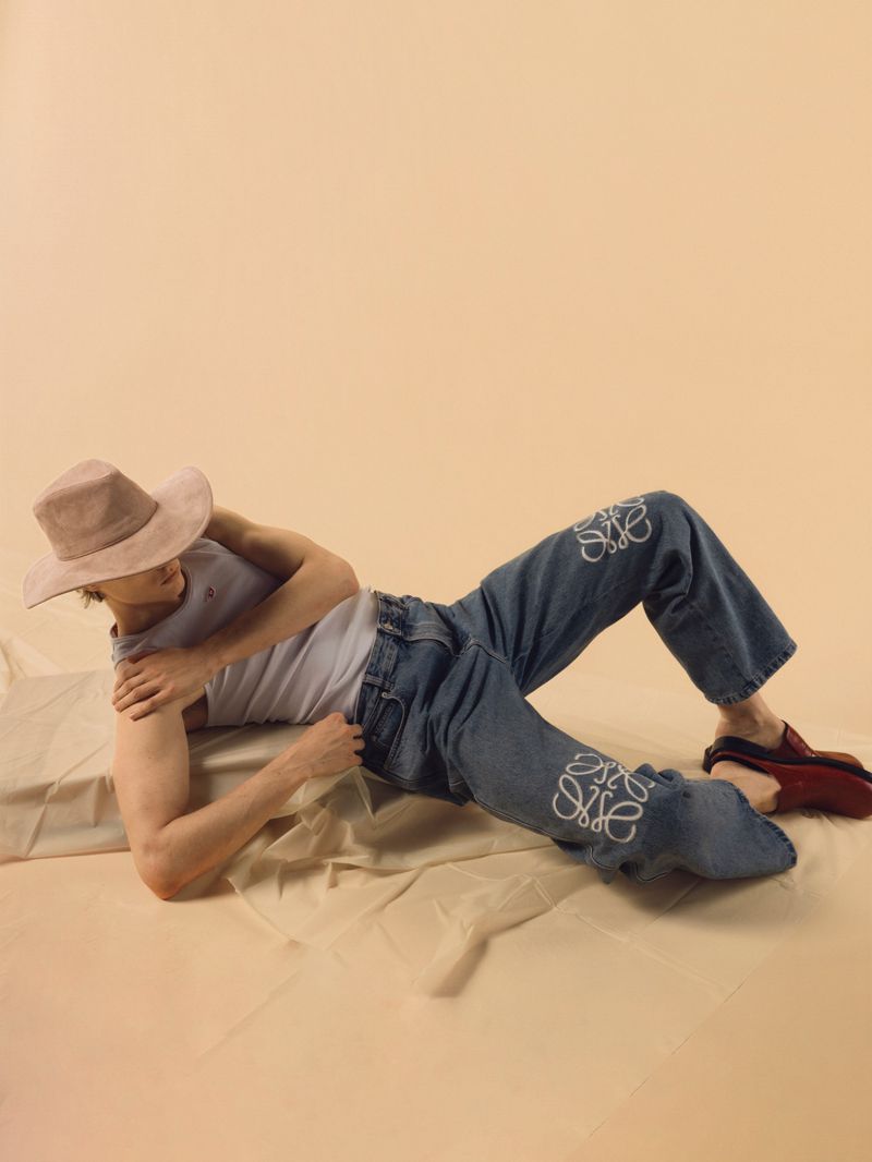 Man in jeans with Loewe logo, tank top, and cowboy hat lies on fabric in a studio setting. Fashion shot, tan background.