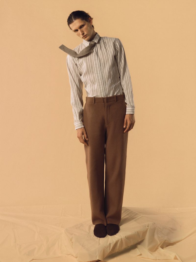A person in a white striped shirt and brown trousers stands on a block covered with a draped cloth in a studio setting.