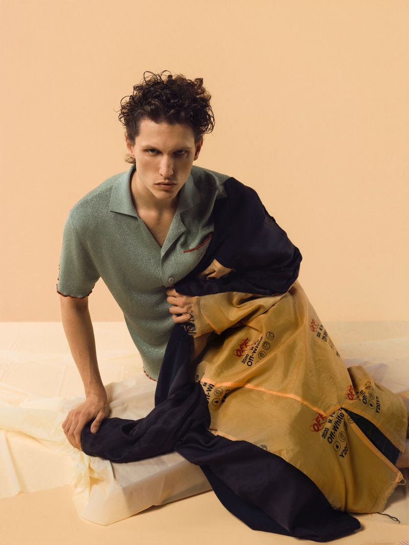 Fashion shot of a young adult male in a green polo shirt, posed in a photo studio setting with clothing props and a light beige backdrop.