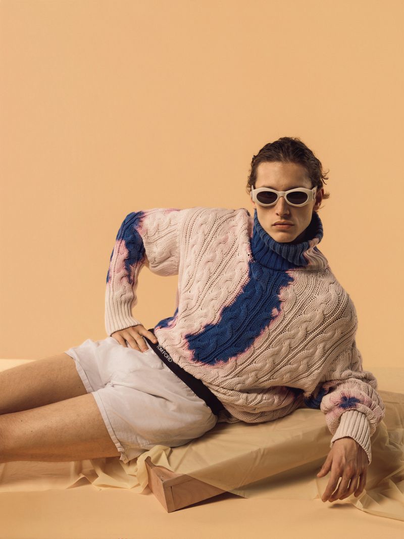 Adult male fashion model in a cream and blue tie dye knit sweater, white shorts, and oval sunglasses, reclining on a beige platform.