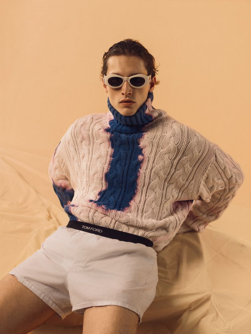 A slender man poses indoors, wearing white sunglasses, a multicolored knit sweater, and light-colored shorts with a dark waistband.