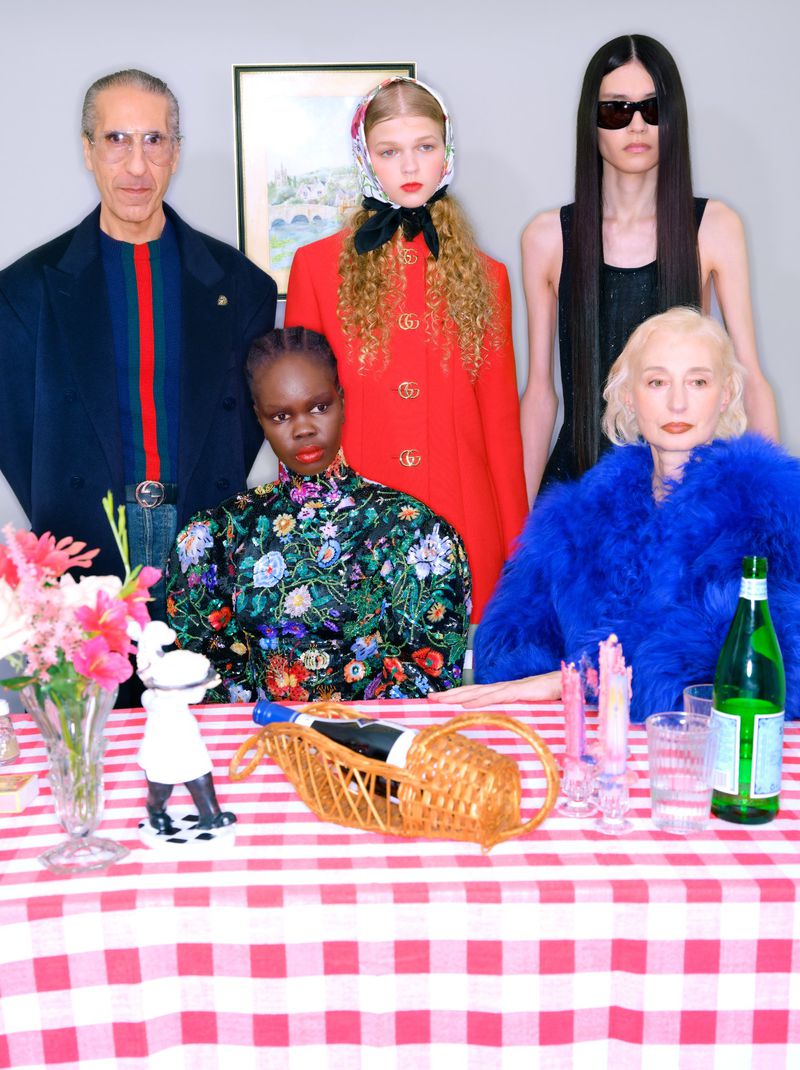 A group of five fashionably dressed adults pose behind a table set with drinks, flowers, and checkered tablecloth, under indoor lighting.