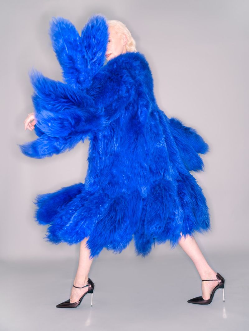A person models a bright blue, textured coat with black, pointed-toe heels against a grey backdrop in a fashion photo studio shot.
