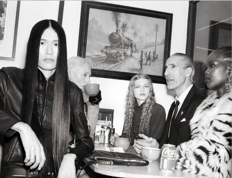 A diverse group of five people are seated at a table inside, with a framed painting of a steam train visible in the background.