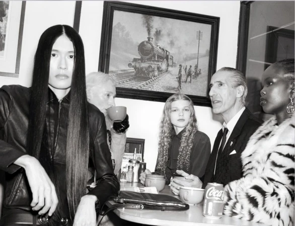 A diverse group of five people are seated at a table inside, with a framed painting of a steam train visible in the background.