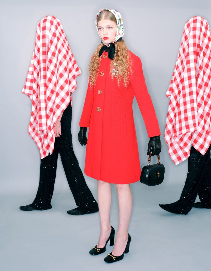 Fashion shot of a woman in a red coat, headscarf, and black heels, flanked by two figures draped in red-and-white checkered tablecloths.