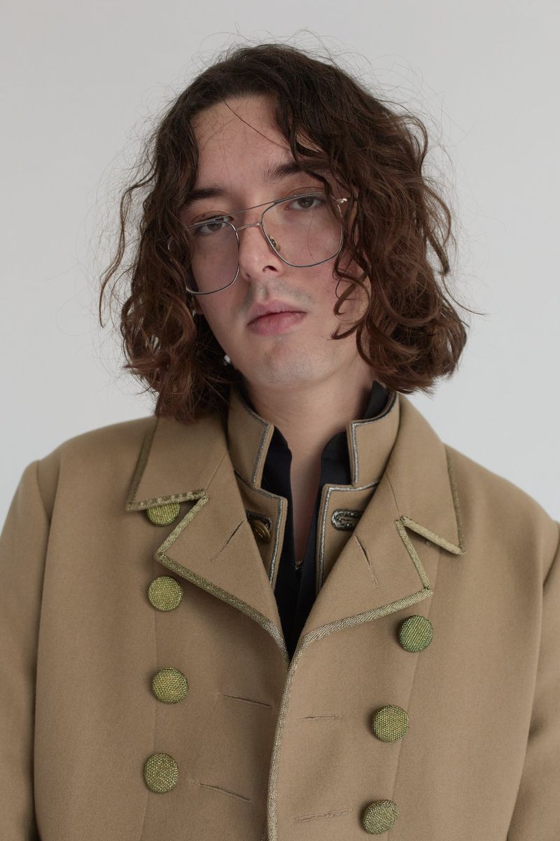 Portrait of a young adult man with long curly hair wearing a tan military-style coat against a neutral white studio backdrop.