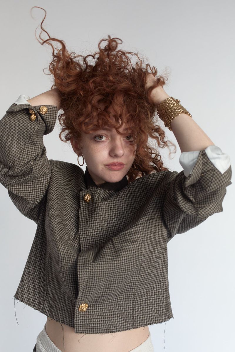 Portrait of a curly-haired young woman in a cropped houndstooth jacket, gold bracelet and hoop earrings, posing indoors against a white backdrop.