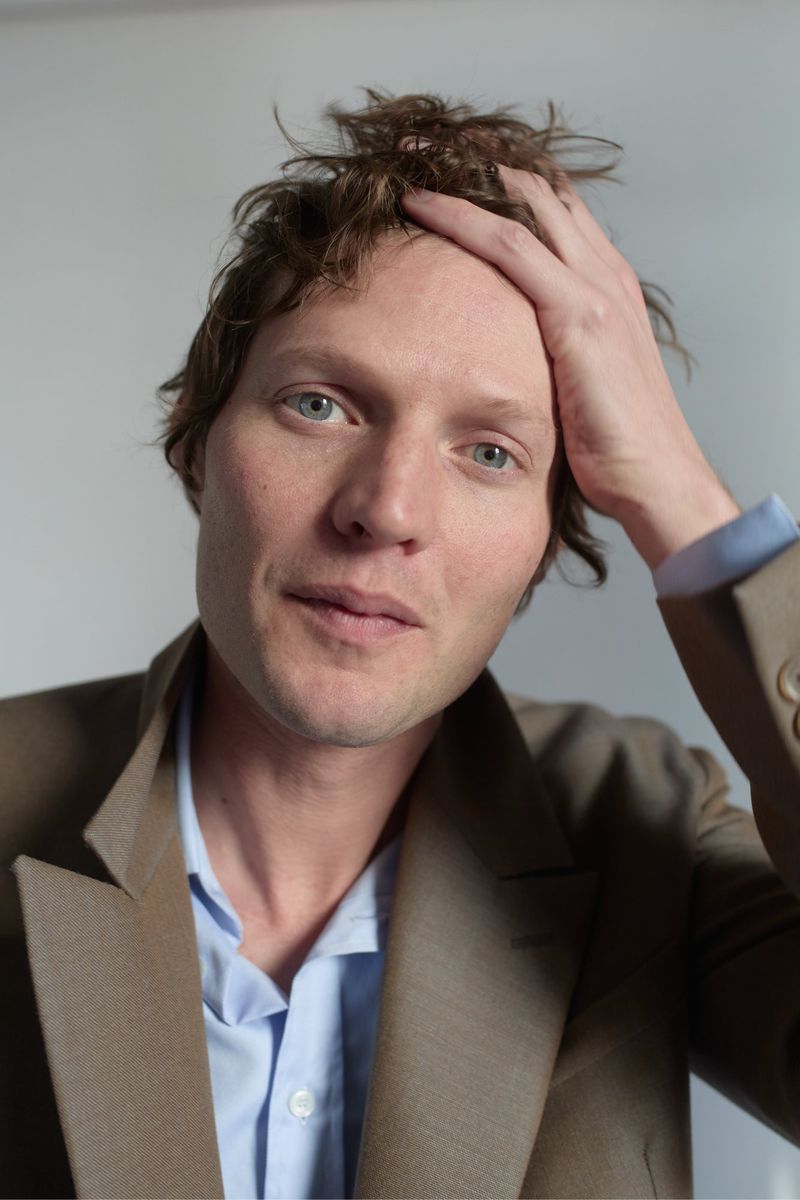 Close-up portrait of a man with his hand on his head. He is wearing a light brown blazer and a blue dress shirt, against a white background.