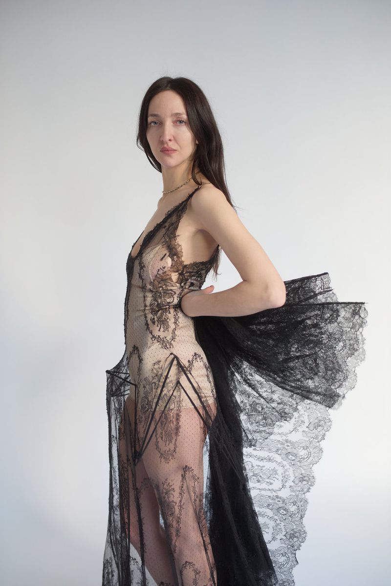 A woman with long dark hair posing in a sheer black lace dress against a plain light gray studio background.