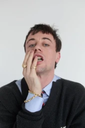 Portrait of a man wearing a dark cardigan over a light blue shirt, with gold rings and a bracelet, touching his face against a white wall.