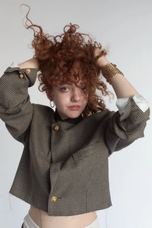 Young woman with curly red hair holding her hands up in her hair, wearing a patterned houndstooth jacket against a plain background.