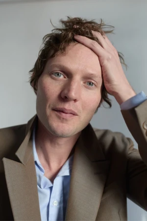 Portrait of a man wearing a brown blazer and blue shirt, with his hand on his head against a plain white wall