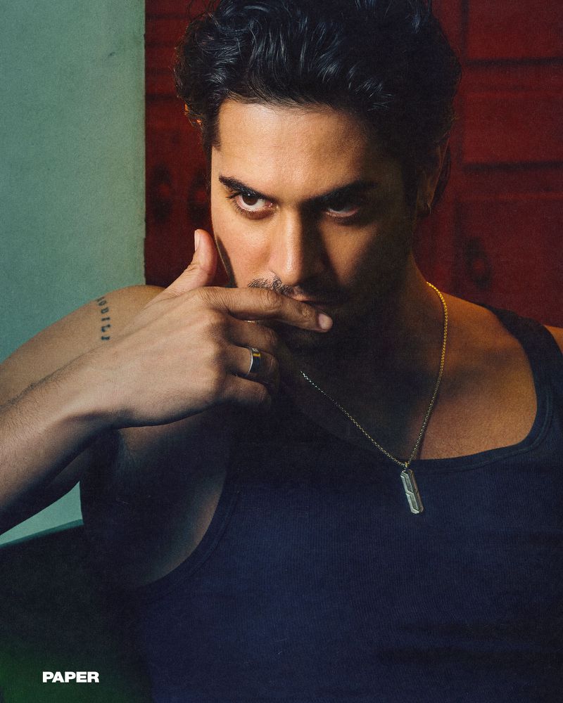 Portrait of a man with dark hair wearing a tank top and gold necklace, set against a textured backdrop of green and red hues.