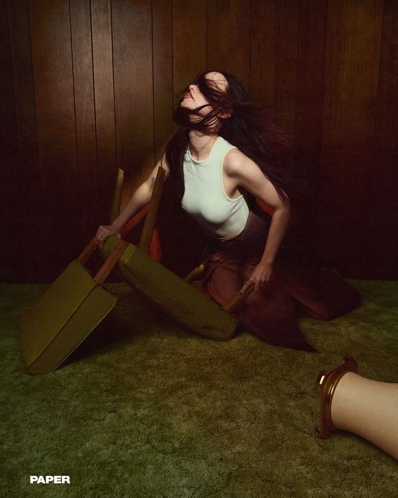 Woman with long dark hair posing in a vintage room with wood paneling, green carpet, and leaning on a chair. 