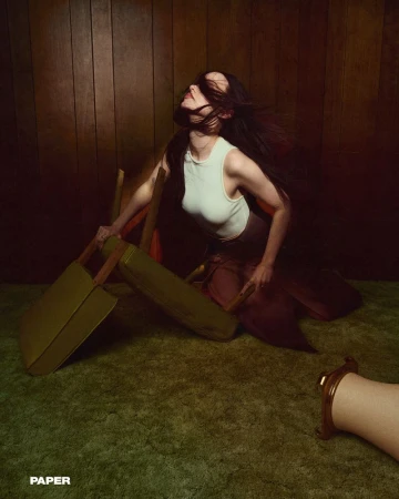 Woman with long dark hair posing in a vintage room with wood paneling, green carpet, and leaning on a chair. 