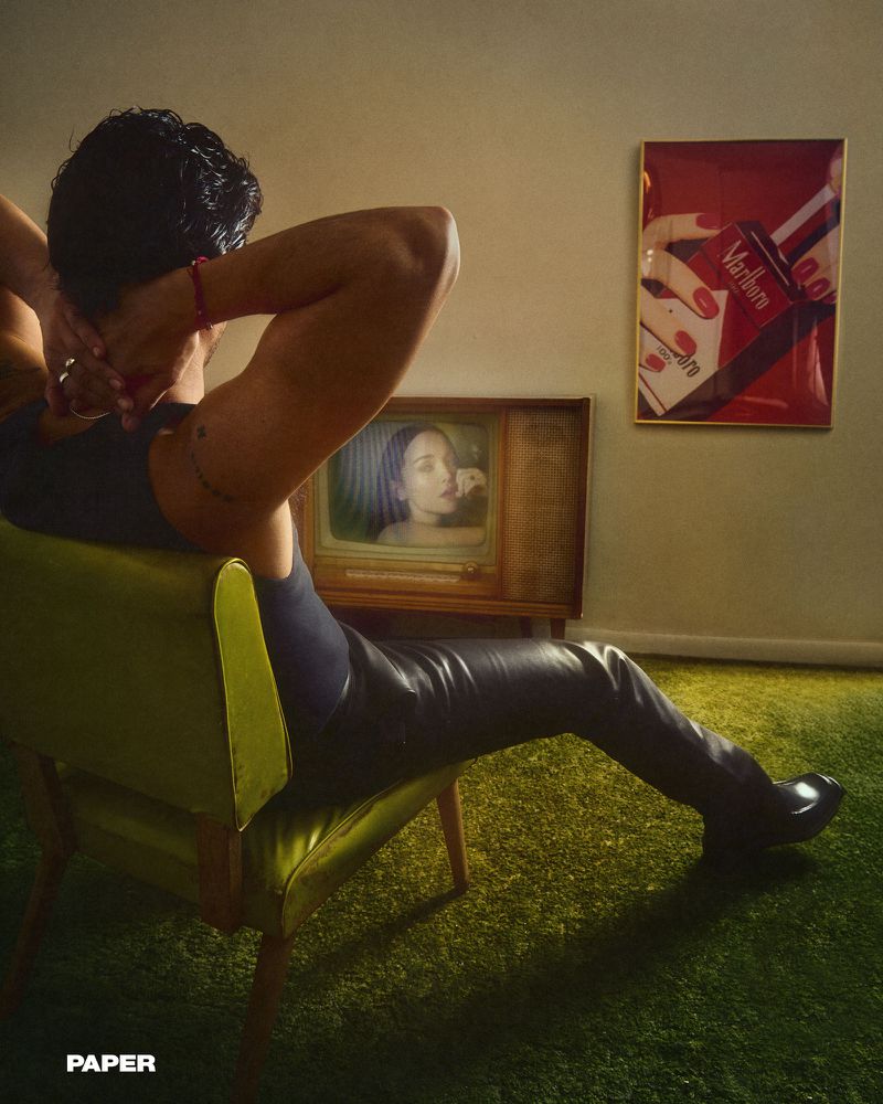 A man lounges in a green chair watching an old TV in a vintage-styled room, with a framed Marlboro ad on the wall.