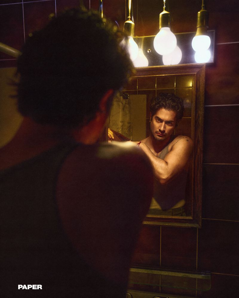 Man gazes into a mirror in a dimly lit bathroom with tiled walls and overhead lights, reflecting a contemplative mood.