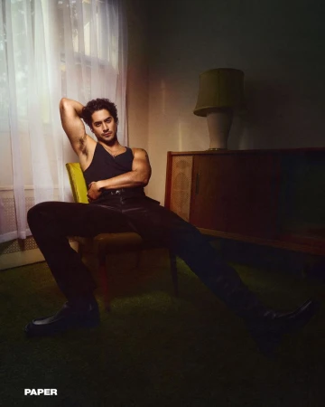 A man in a black tank top and pants poses in a yellow chair with a vintage cabinet and lamp in the background, creating a dramatic portrait.