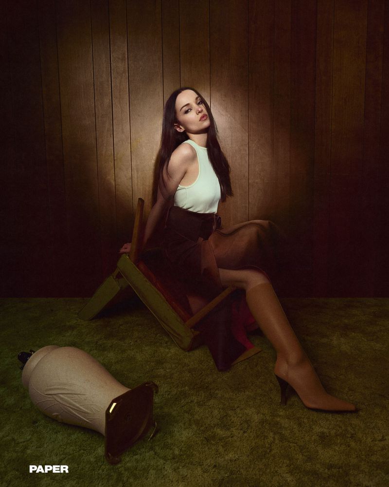 A brunette woman poses on a lounge chair in tall boots and a skirt in a warm-toned studio with a wooden wall.