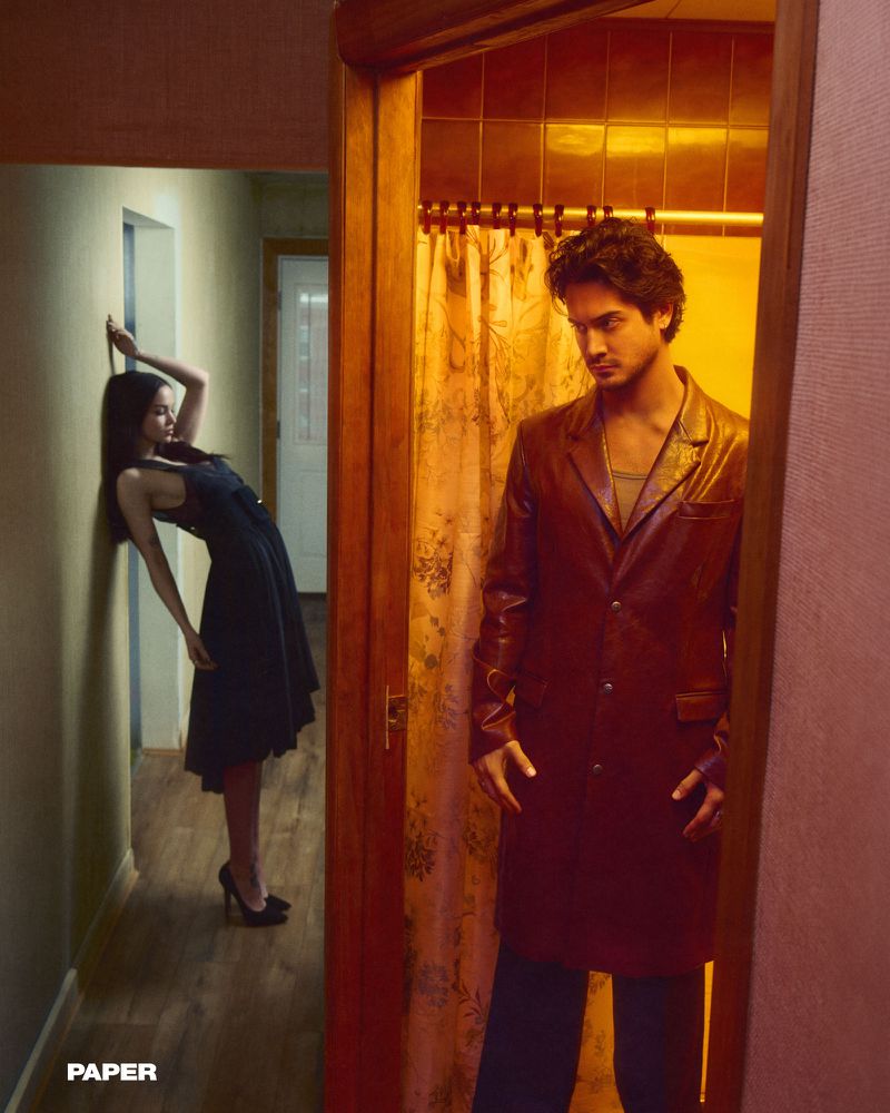 Fashion editorial shot of two people in a hallway, a man in a long leather jacket stands in a doorway as a woman poses in the background.