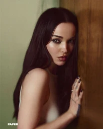 Portrait of Dove Cameron with dark hair leaning against a wooden door for Paper Magazine