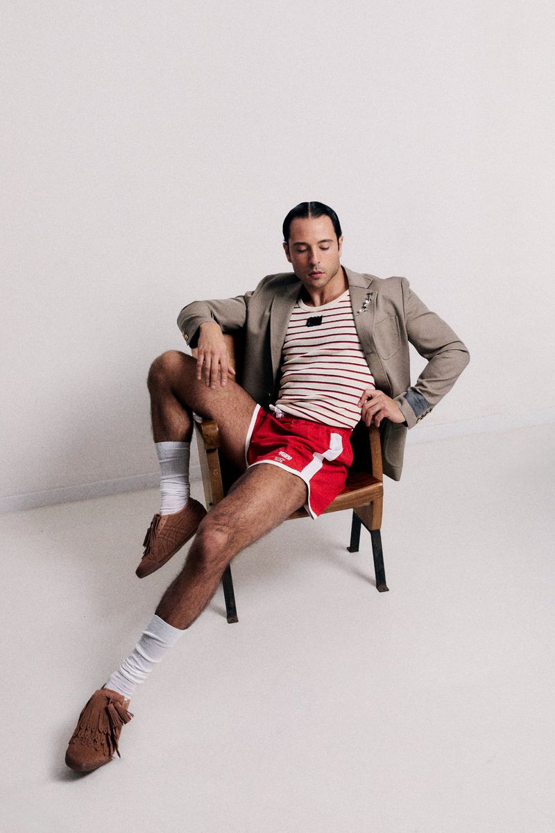 Adult male sitting in a chair wearing a striped shirt, blazer, red shorts, brown shoes, and white leg warmers in a light studio.