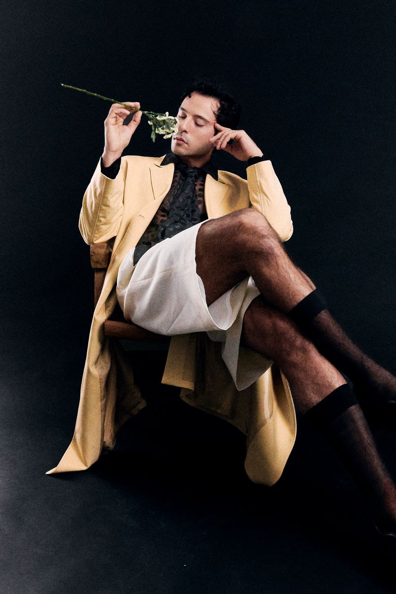 Young adult male fashion portrait sitting in wooden chair holding a rose, wearing a yellow trench coat and white shorts on black background.