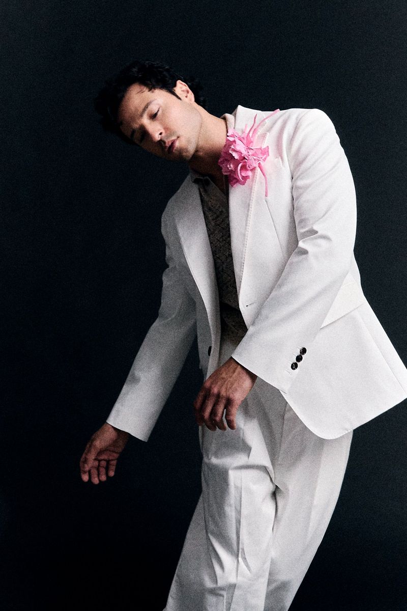 Male model in white suit with pink flower poses against dark background in a fashion portrait. Studio shot, eye-level angle.