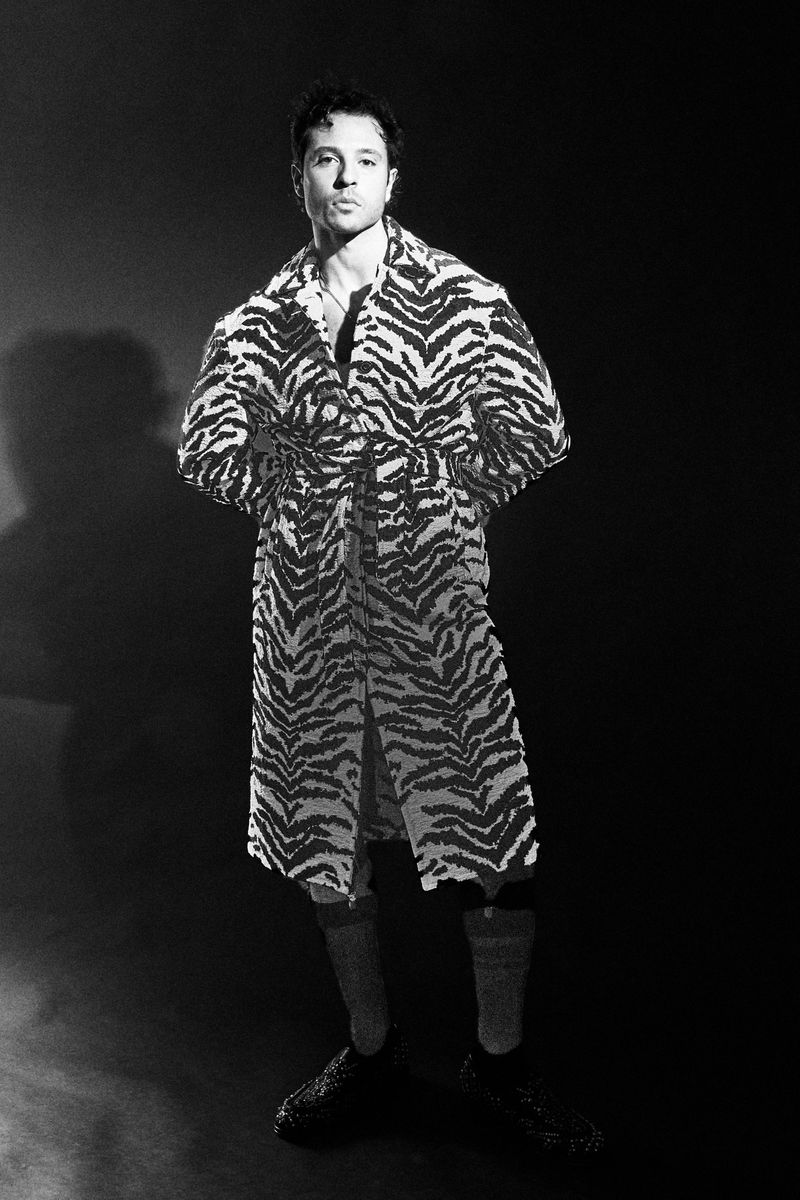 Black and white portrait of a man wearing a tiger-striped robe, socks and shoes, posing in front of a dark, hazy background.