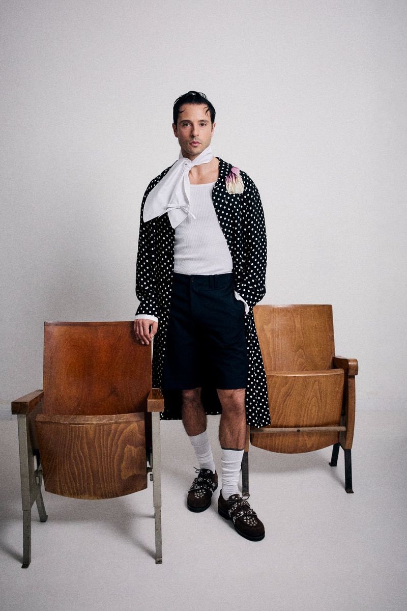 A man posing in a polka dot robe, white shirt, and dark shorts stands next to two wooden chairs against a white backdrop in a studio.