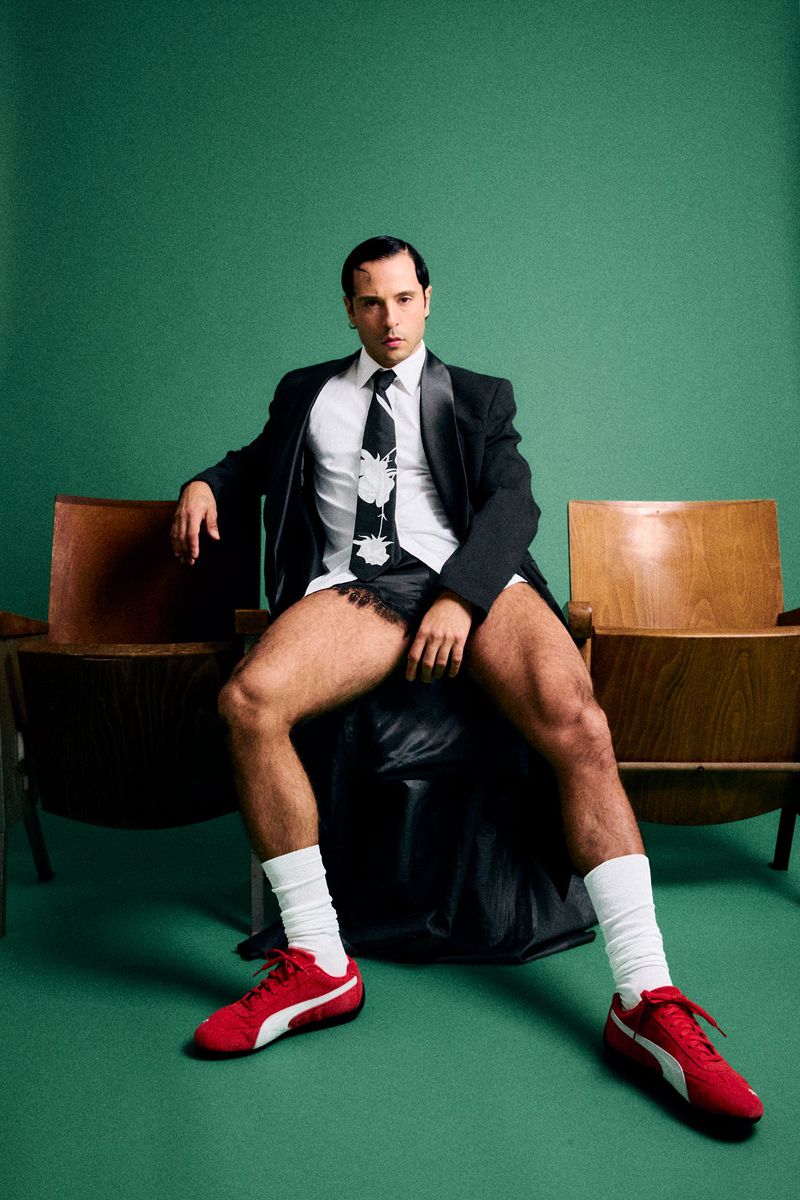 Stylish man in a suit jacket, patterned tie, and underwear, paired with red sneakers and white socks, poses in a studio setting.