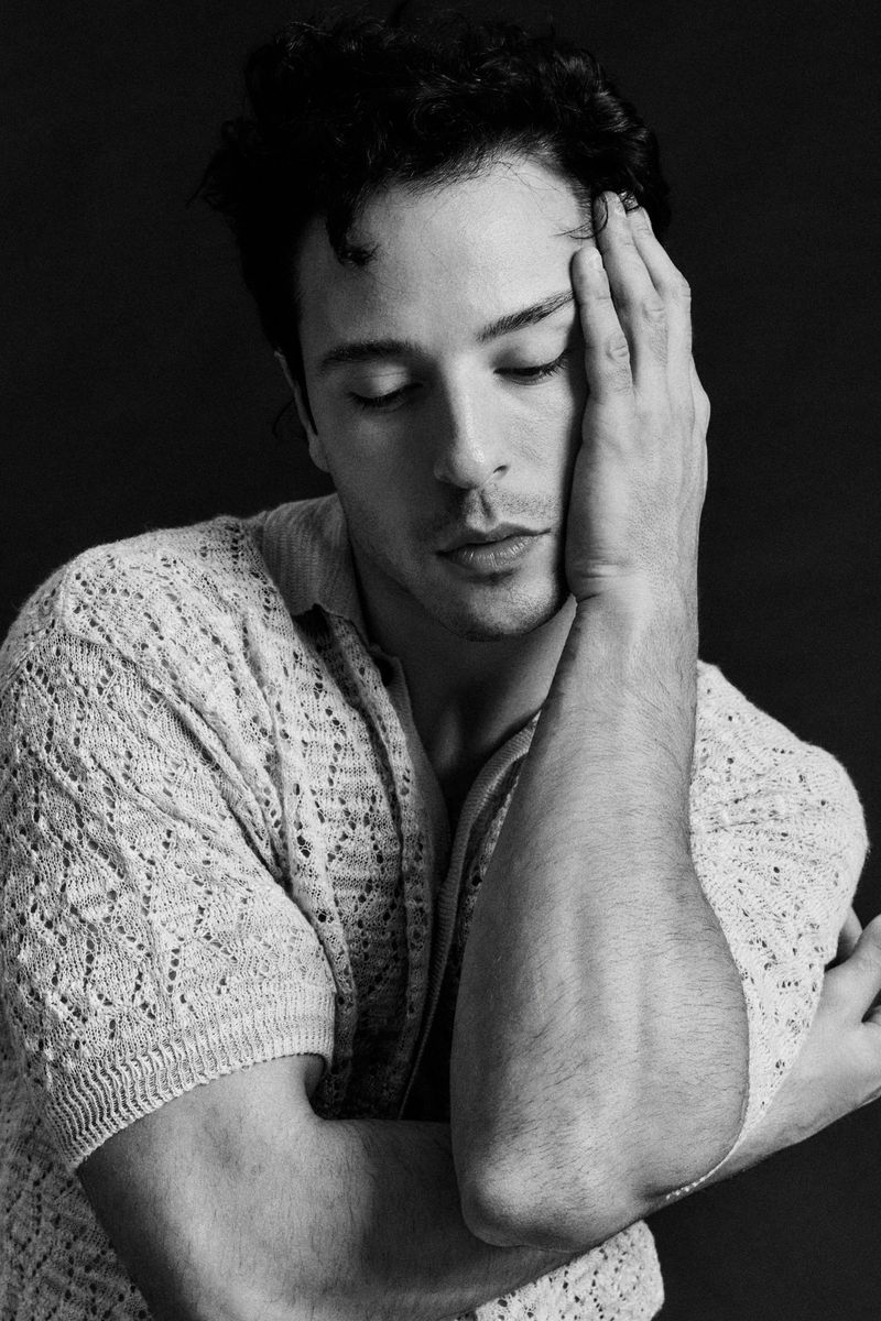 Black and white portrait of an adult male with dark hair, eyes closed, wearing a knitted sweater, with his hand on his head in studio lighting.