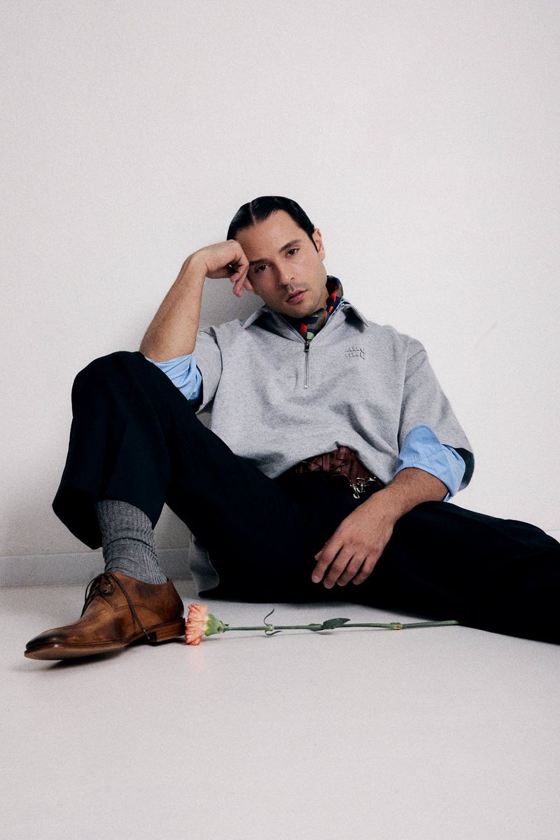 Stylish man sits against a white wall, wearing a grey sweatshirt, dark trousers, and brown leather shoes, holding a pink carnation.