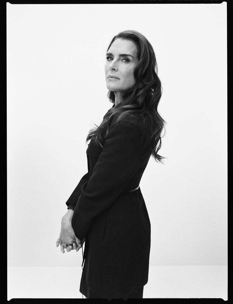 Black and white portrait of Brooke Shields, side view, wearing a dark jacket with a belt, long wavy hair, and hands clasped in front.