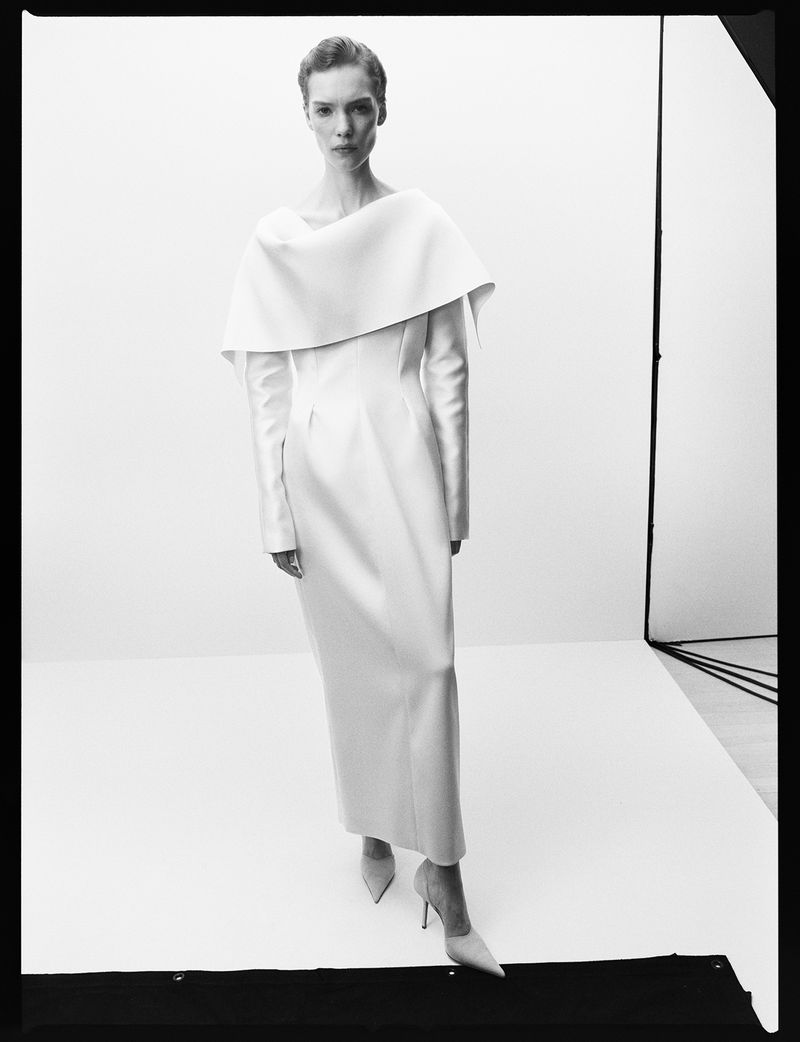 Elegant woman in long-sleeved white gown and high heels stands in a photo studio with white backdrop and minimalist, modern design.