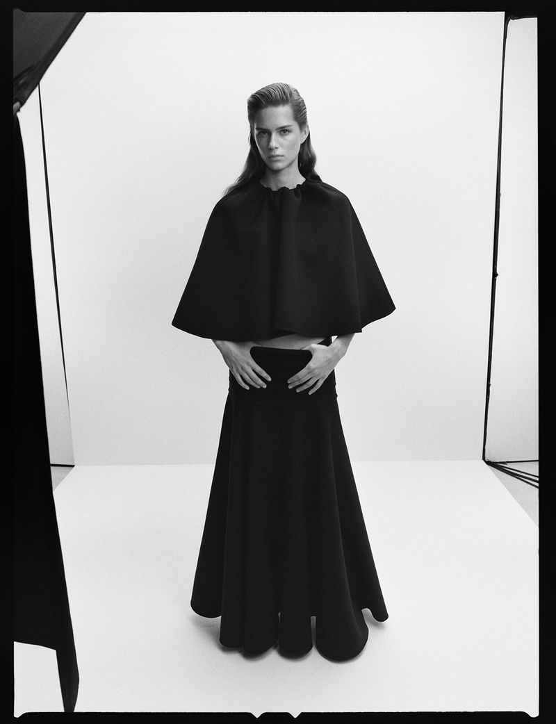 Black-and-white studio shot of a model in a black cape top and long black skirt, hands resting on her waist, gazing at the camera.