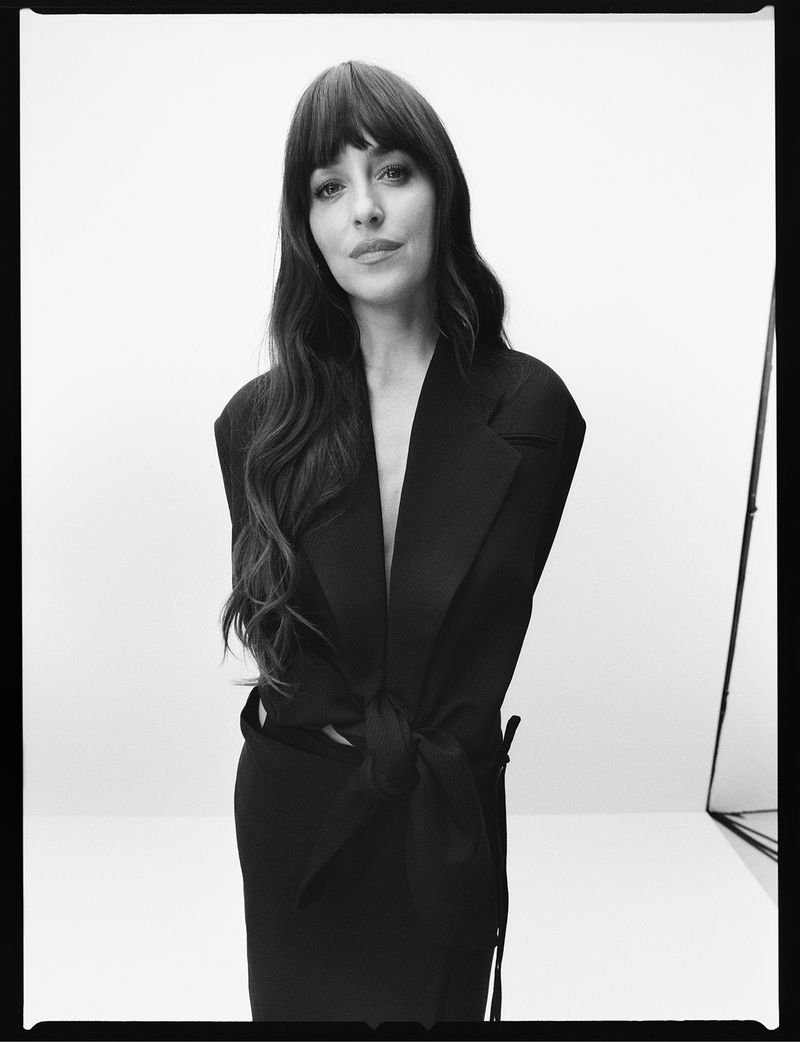 Black and white portrait of a woman with long hair and bangs, wearing a dress with a tie belt, posing against a white backdrop.