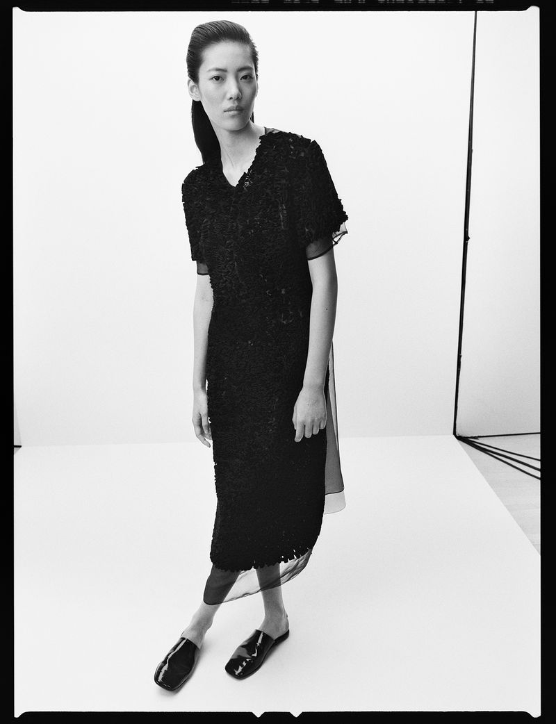 Studio shot of an adult female model wearing a black dress and patent leather slide shoes against a white backdrop with studio lighting.
