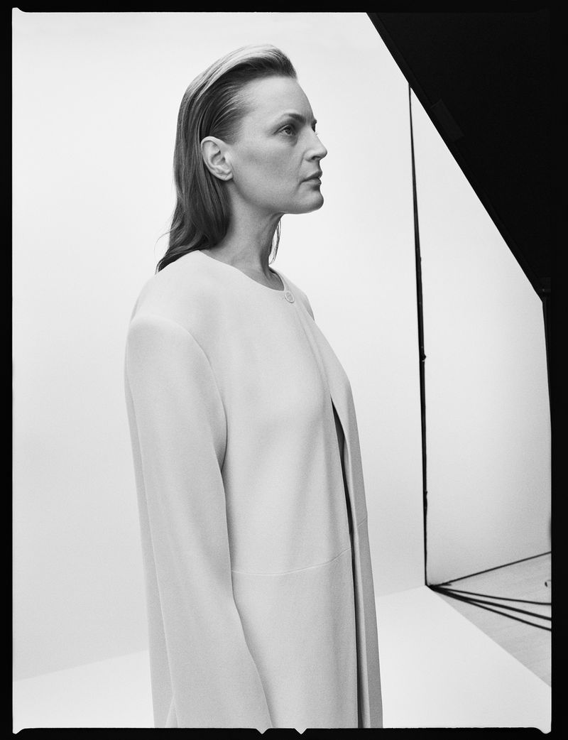 Monochrome portrait of an adult woman in a light-colored coat posing in a studio with soft lighting and a simple background.