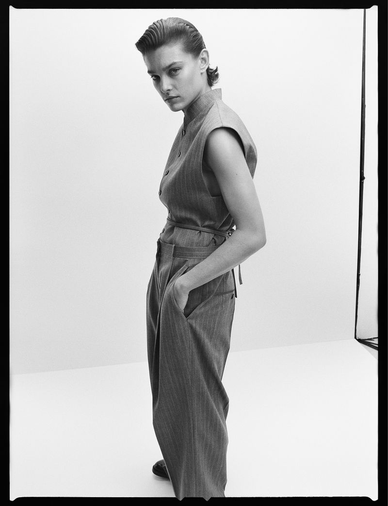 Model in sleeveless top and trousers with slicked-back hair posing against a white studio sweep background